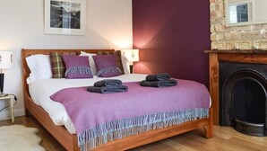A bedroom with a wooden bed, purple bedding, a fireplace, and a stone wall. - Brae Cottage (Crieff)