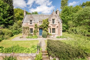 A stone house with a steeply pitched roof, a central door, and a well-maintained garden. - Gardeners Cottage (Crieff)