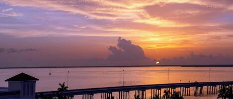 A sunset over a calm body of water with a bridge and palm trees in the foreground.