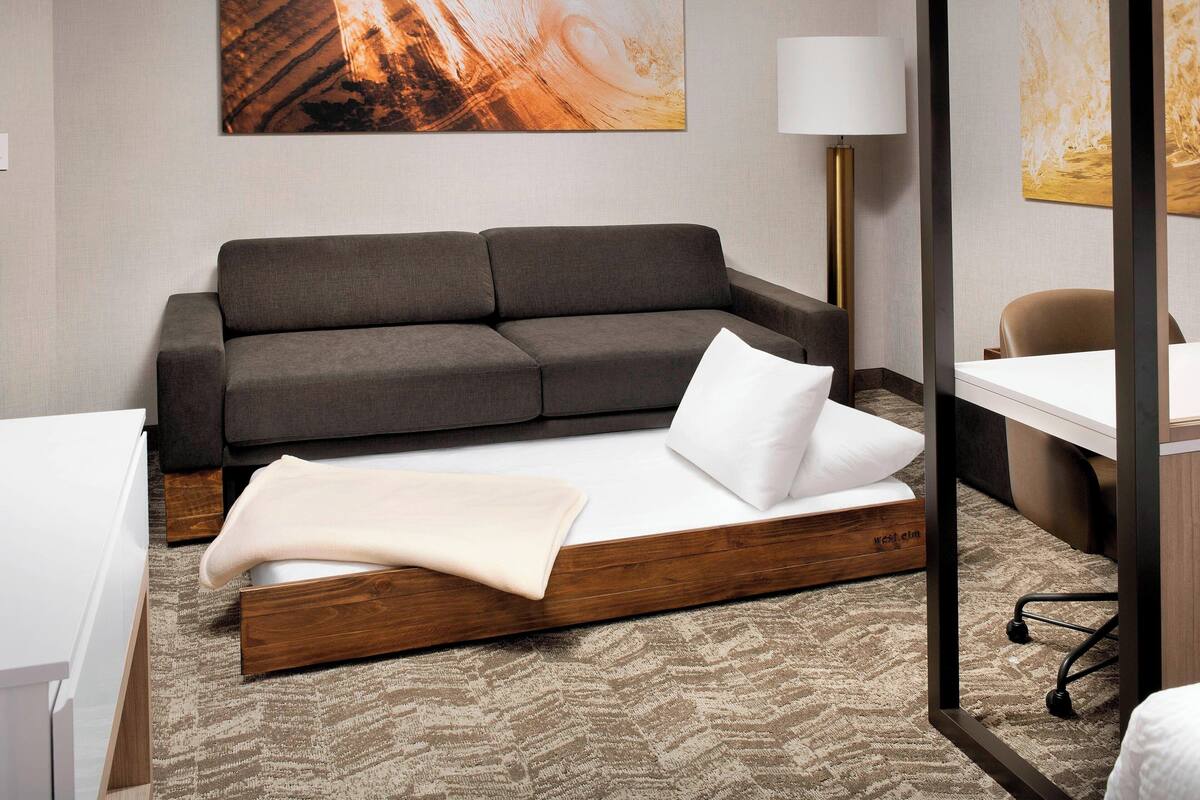A modern hotel room with a grey sofa, a bed with a wooden frame, a desk, and a lamp.