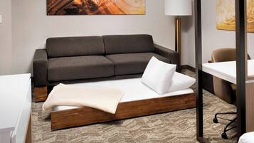 Una habitación de hotel moderna con un sofá gris, una cama con estructura de madera, un escritorio y una lámpara.