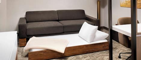 A modern hotel room with a grey sofa, a bed with a wooden frame, a desk, and a lamp.