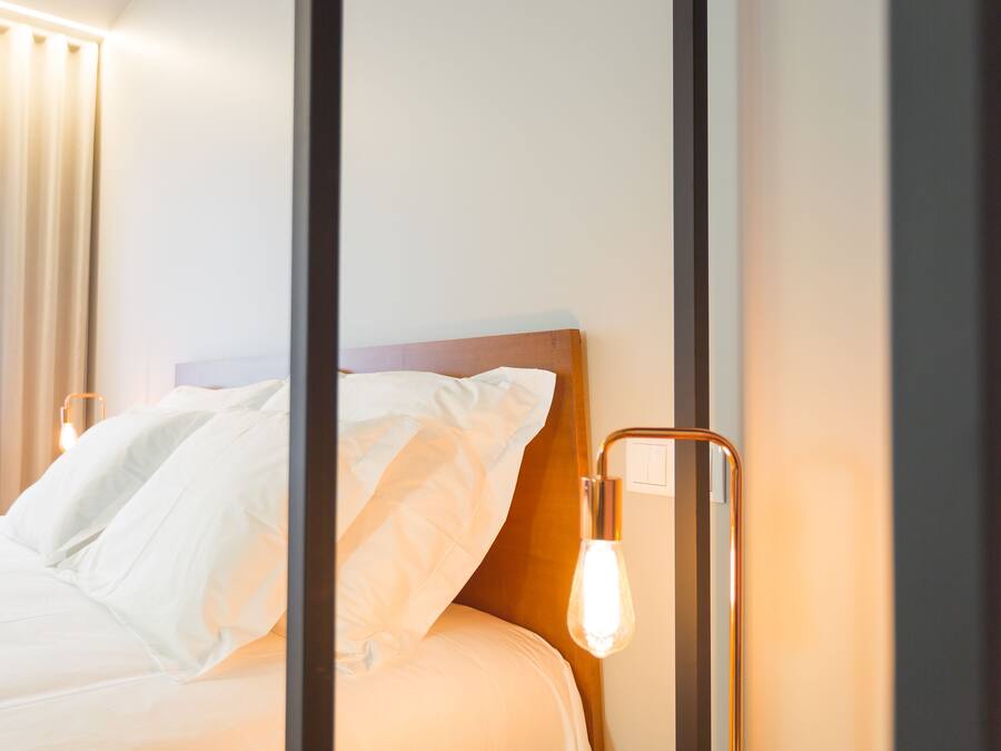 Una cama bien tendida con sábanas blancas, cabecera de madera y dos lámparas fijadas a la pared.