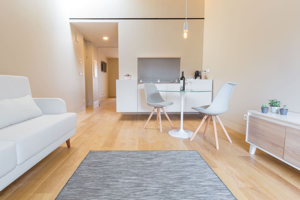 A modern living room with a white sofa, a dining table with two chairs, and a kitchen area in the background.