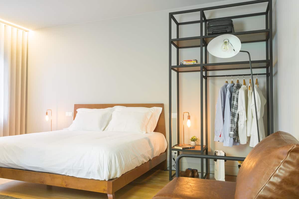 A neatly made bed with white linens, a wooden headboard, a black metal shelf with clothes and a lamp, and a leather chair.