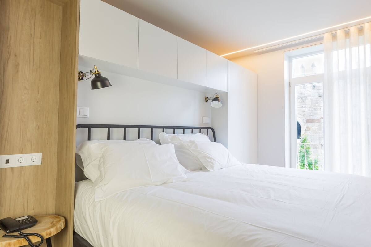 A neatly made bed in a hotel room with a wooden headboard, a bedside telephone, and a window with curtains.