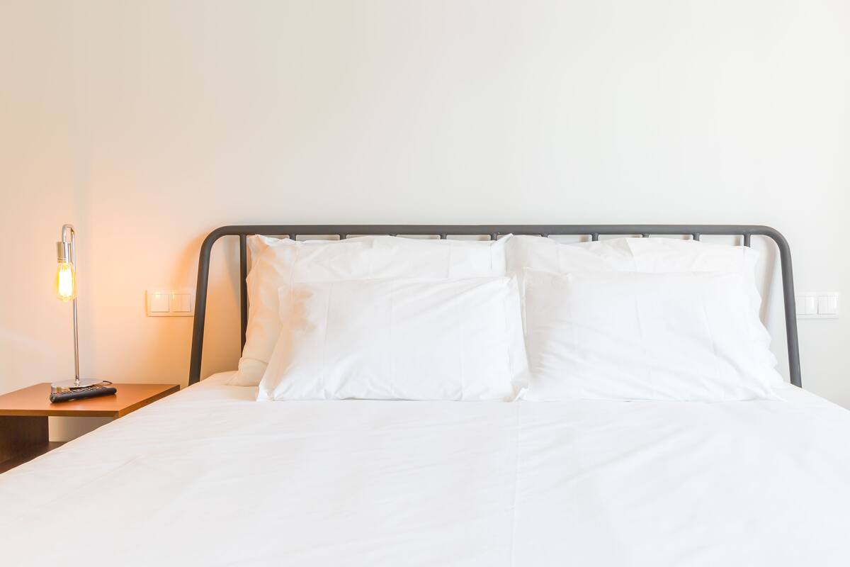 A neatly made bed with white linens and a metal headboard. There is a bedside table with a lamp and a remote control.
