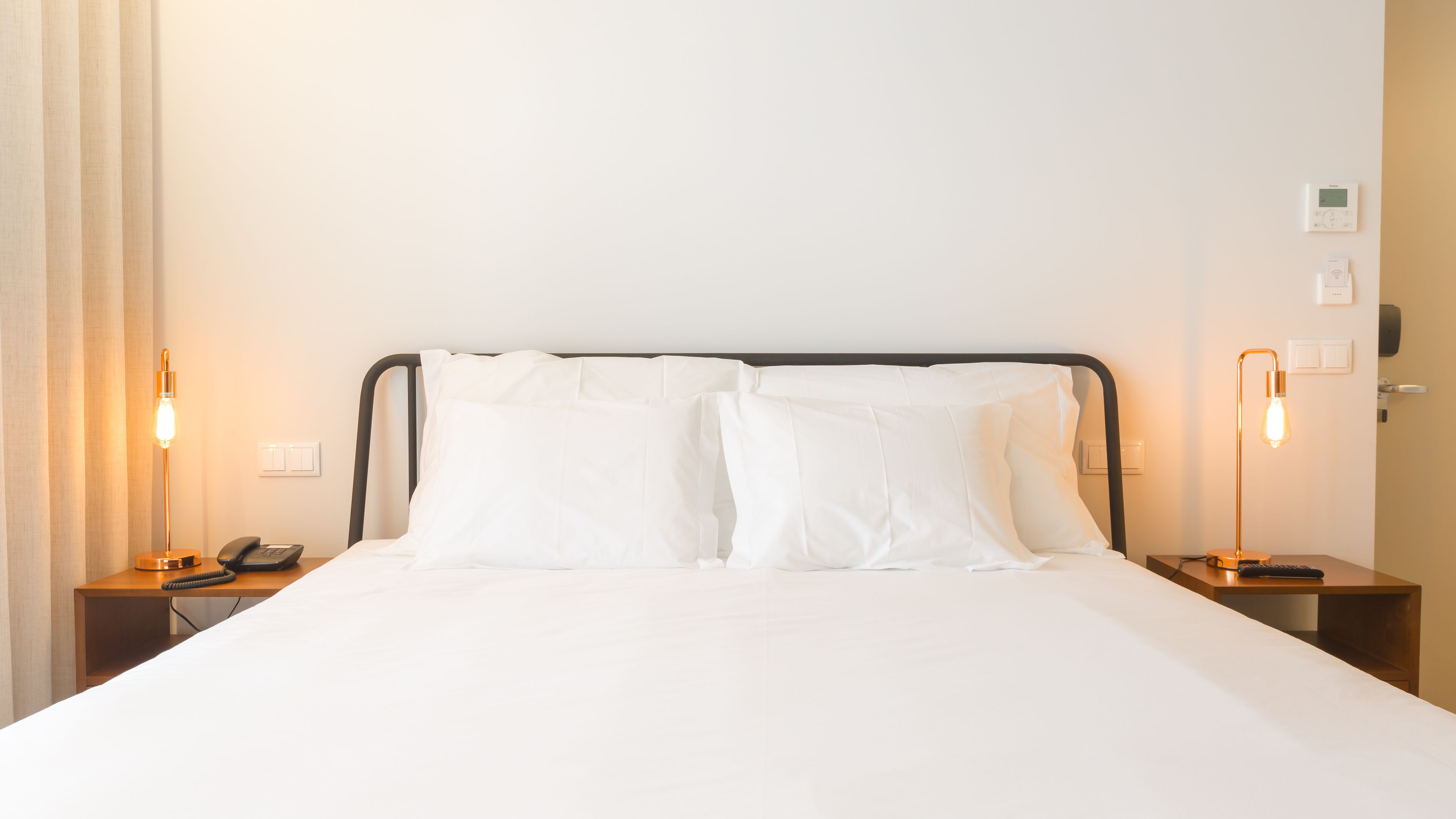 A hotel room with a bed, two bedside tables, a lamp, a phone, and a thermostat on the wall.