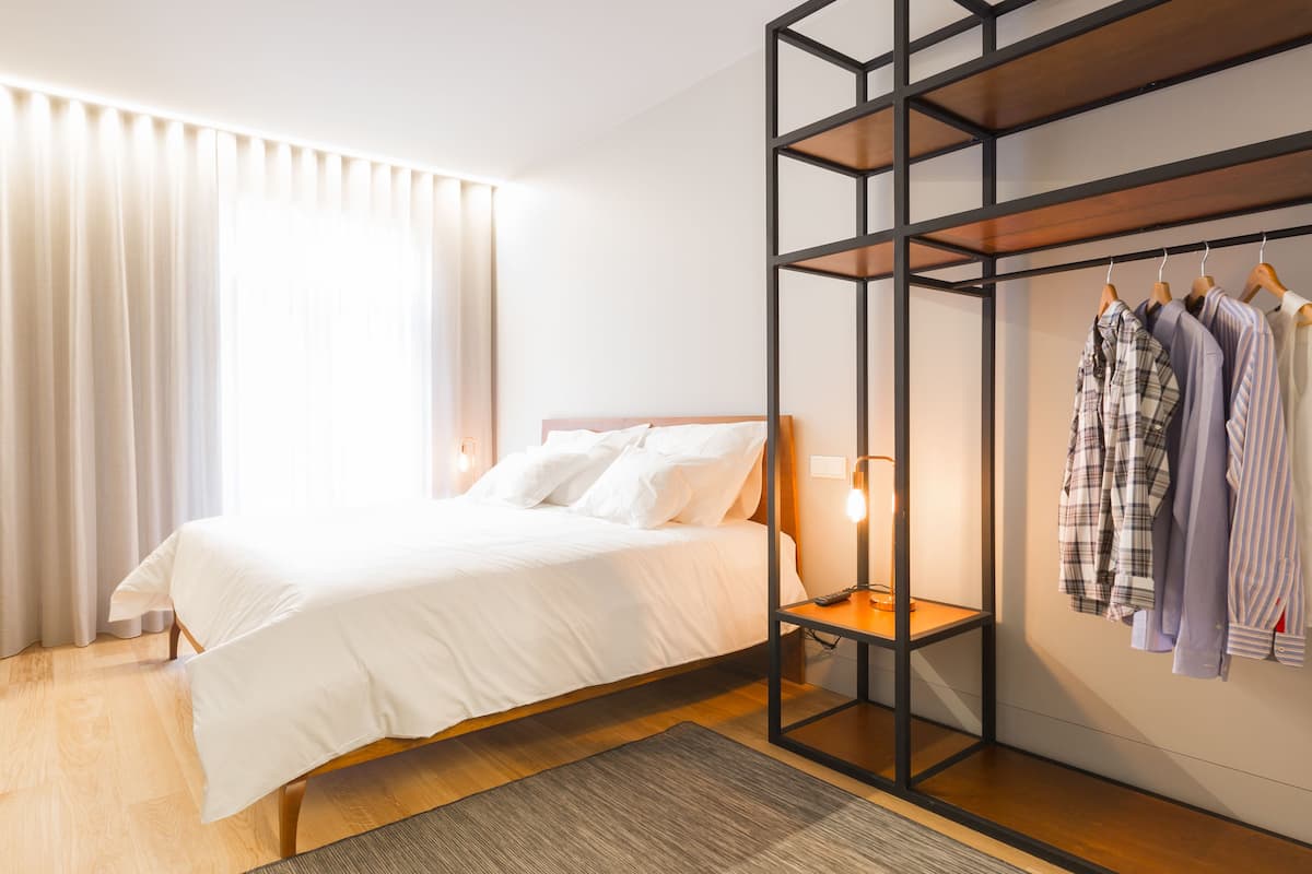 A modern hotel room with a large bed, a wooden floor, a desk, a chair, a wardrobe with clothes, and a floor lamp.