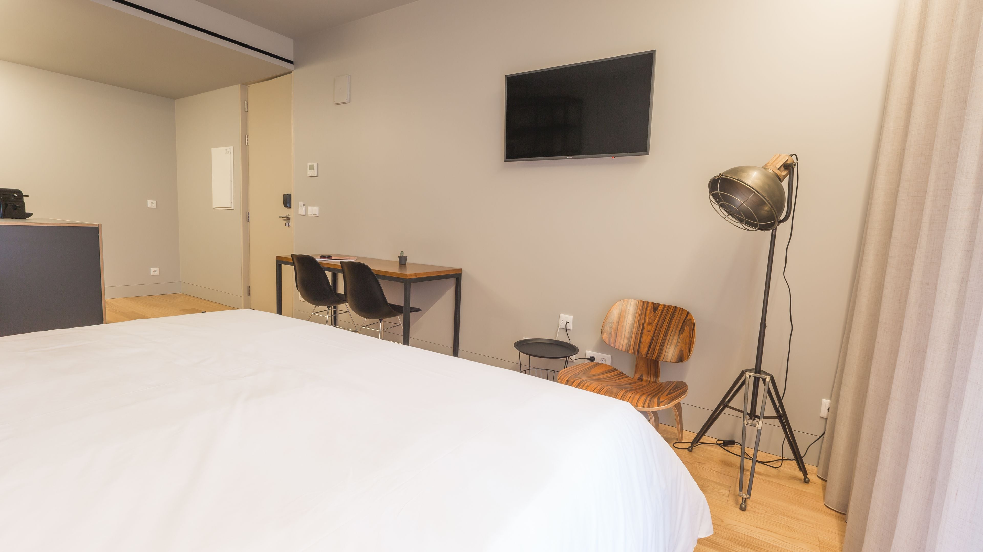A hotel room with a bed, a desk, two chairs, a television, a floor lamp, and a small table with a plant.