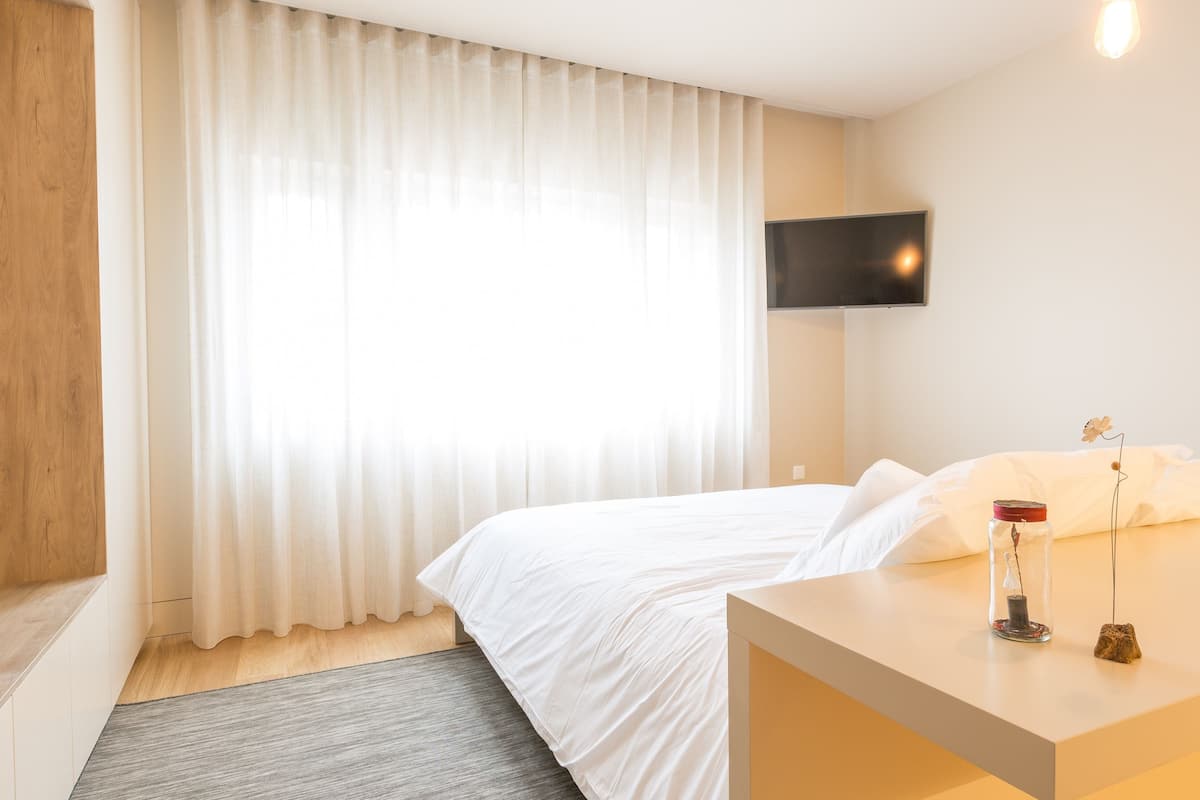 A hotel room with a bed, a TV mounted on the wall, a bedside table with a vase and a candle, and a window with sheer curtains.