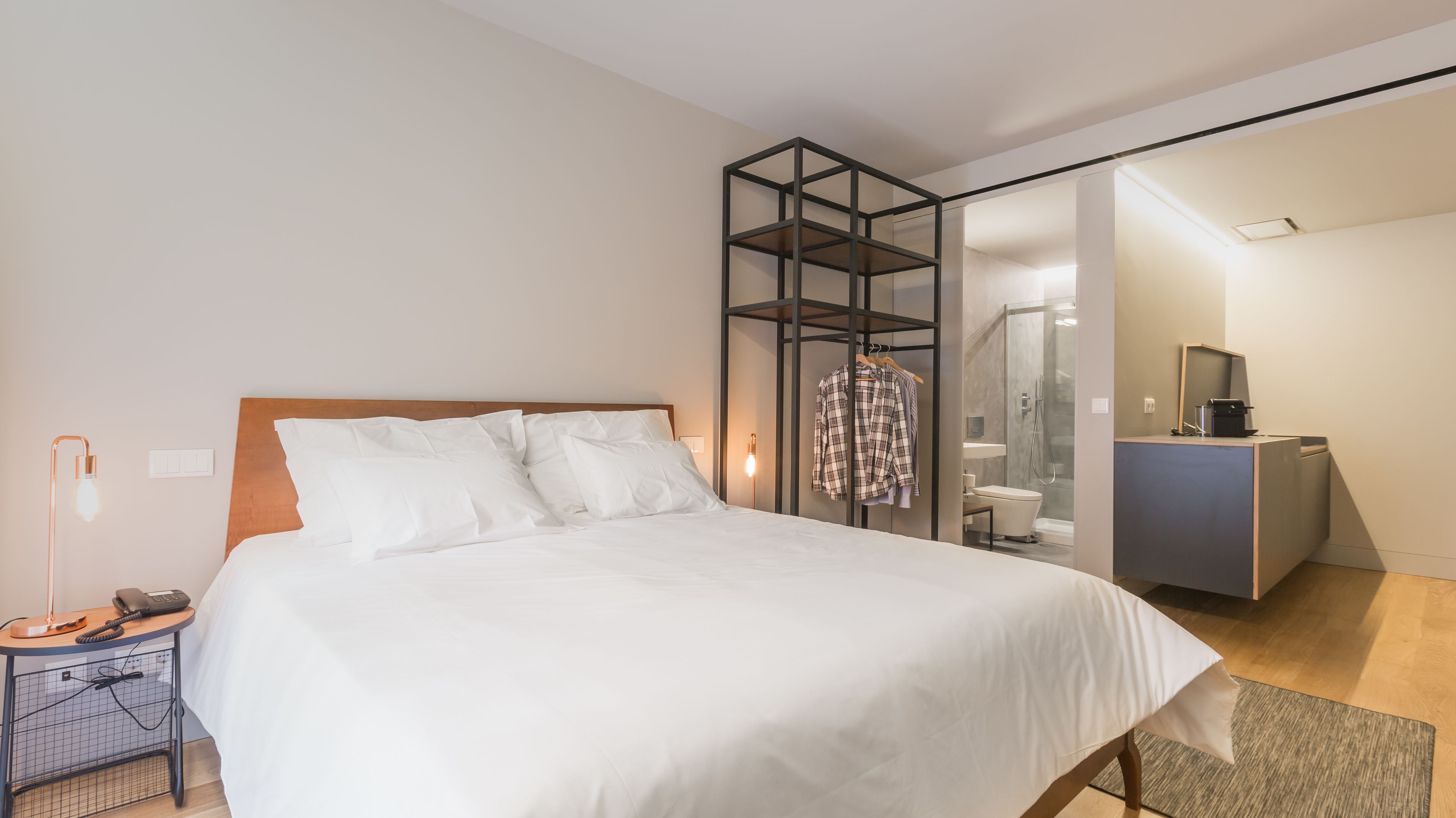 A modern hotel room with a large bed, a bedside table with a lamp, a wardrobe, and a bathroom visible through a glass partition.