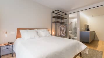 A modern hotel room with a large bed, a bedside table with a lamp, a wardrobe, and a bathroom visible through a glass partition.