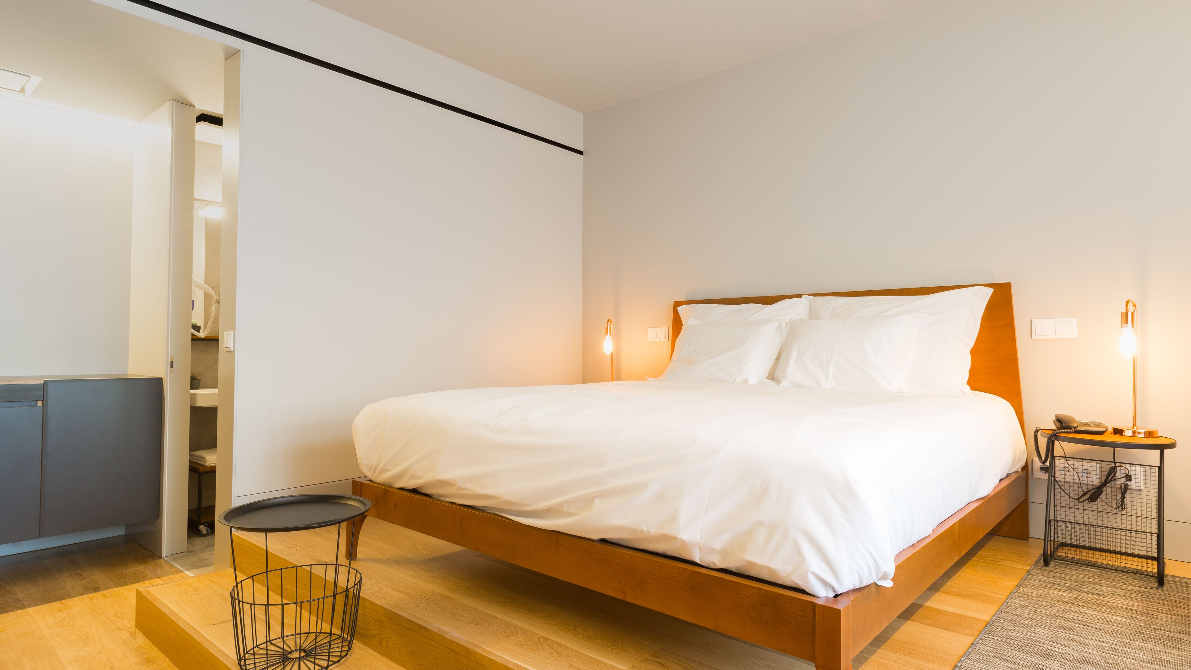 A modern hotel room with a wooden bed, a small round table, a bedside lamp, and a desk with a chair.