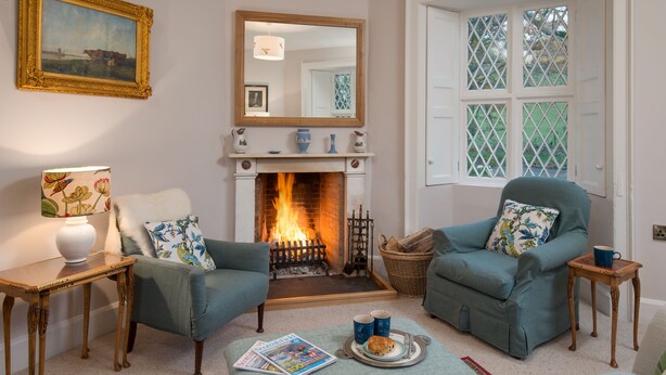 A cozy living room with a fireplace, two armchairs, a coffee table with a magazine, and a window with lattice. - East Gatehouse Lodge (Crieff)