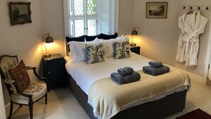 A neatly arranged bedroom with a large bed, two bedside tables with lamps, a chair, a window with a view, and a robe rack with robes. - East Gatehouse Lodge (Crieff)