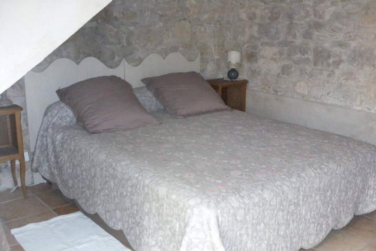 Une chambre avec un mur en pierre, un lit recouvert d’une couvre-lit à motifs, deux oreillers, une table de chevet avec une lampe et un tapis blanc sur le sol.