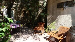 A shaded outdoor seating area with wicker chairs and a wooden table, surrounded by greenery and stone walls.
