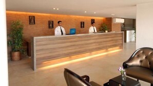 Un mostrador de recepción con dos empleados, un mostrador de madera, plantas en macetas y un sofá de cuero.