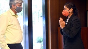 Dos personas de pie frente a una puerta, una vestida con una camisa amarilla y la otra con un traje negro.