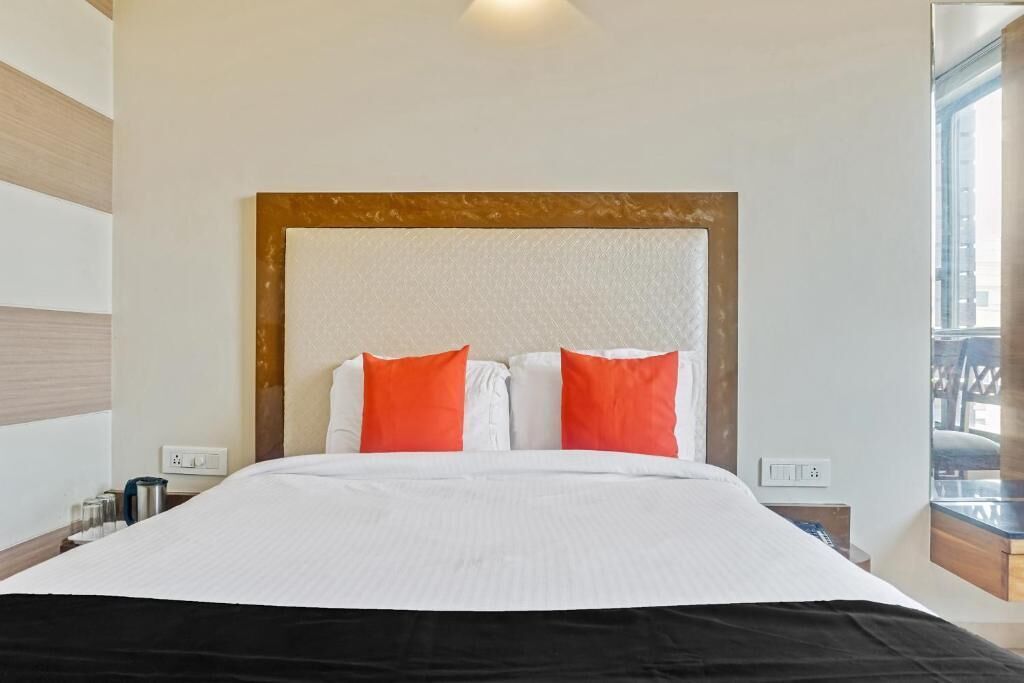 A hotel room with a large bed, white bedding, and orange pillows. There are two bedside tables with lamps and a window with a view.