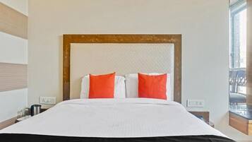 A hotel room with a large bed, white bedding, and orange pillows. There are two bedside tables with lamps and a window with a view.