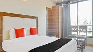 Habitación de hotel con una cama grande, cabecero de madera, balcón con vistas y una pequeña mesa con sillas.