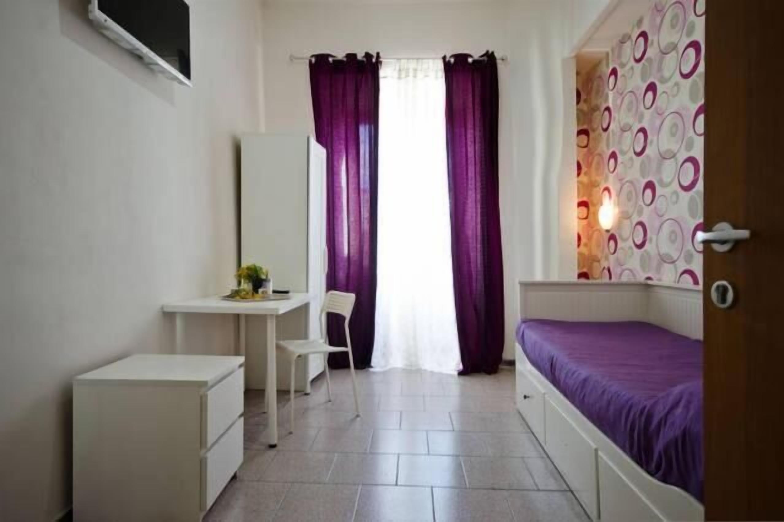 A small, well-lit room with a bed, a desk, and a chair.