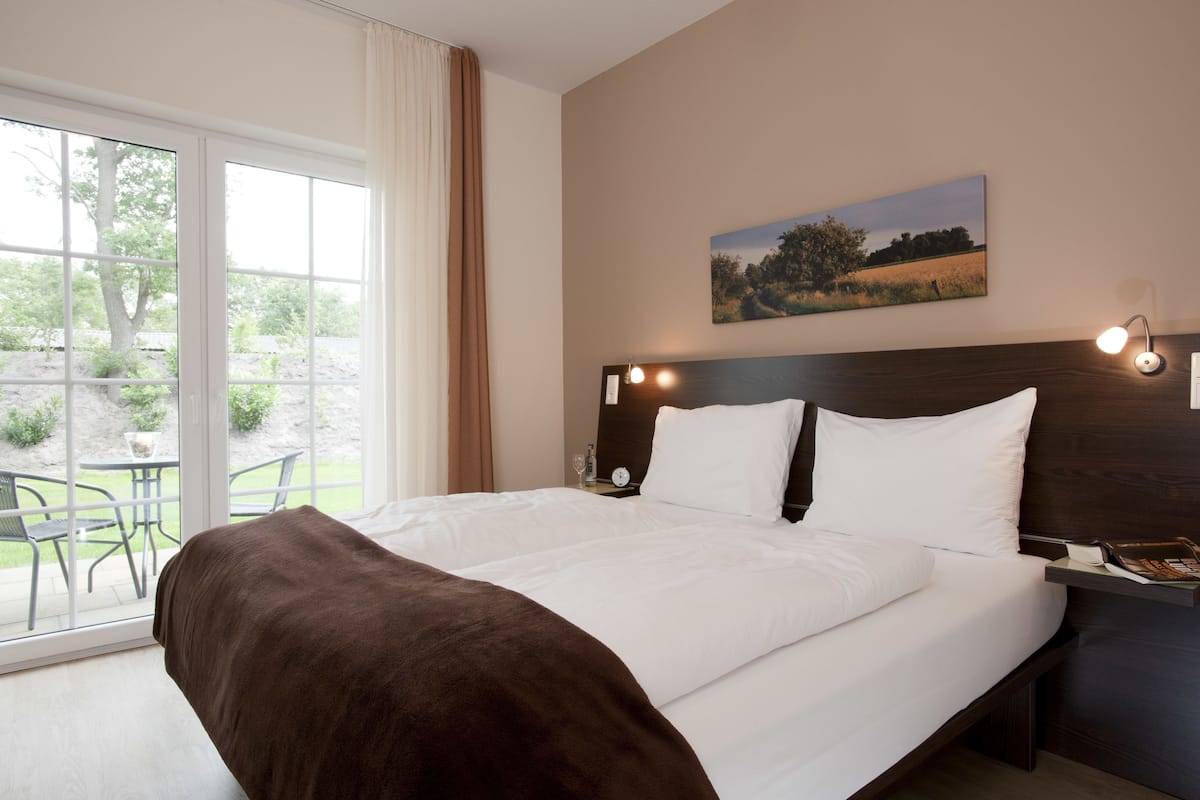 A hotel room with a large bed, a window with a view of a garden, a painting on the wall, and a bedside table with a lamp.
