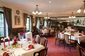 A dining area with multiple tables set for a meal, featuring chandeliers and a view of the kitchen in the background. - Hotel Meiners (Hatten)