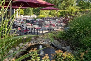 An outdoor patio with tables and chairs, a small water feature, and surrounding greenery. - Hotel Meiners (Hatten)