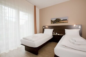 A hotel room with two single beds, a wooden headboard, a bedside table with a lamp, and a painting on the wall. - Hotel Meiners (Hatten)