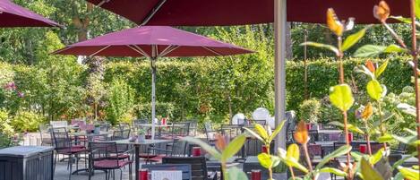 An outdoor seating area with tables and chairs under red umbrellas, surrounded by greenery.