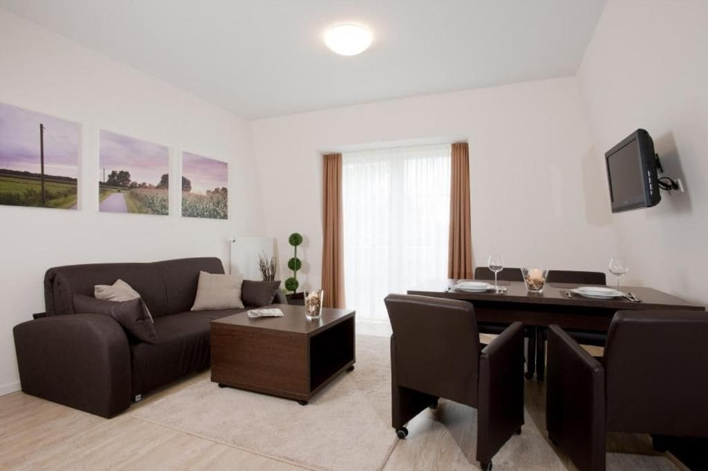 A modern living room with a sofa, coffee table, and dining area. There are three framed pictures on the wall, a flat-screen TV mounted on the wall, and a window with curtains.