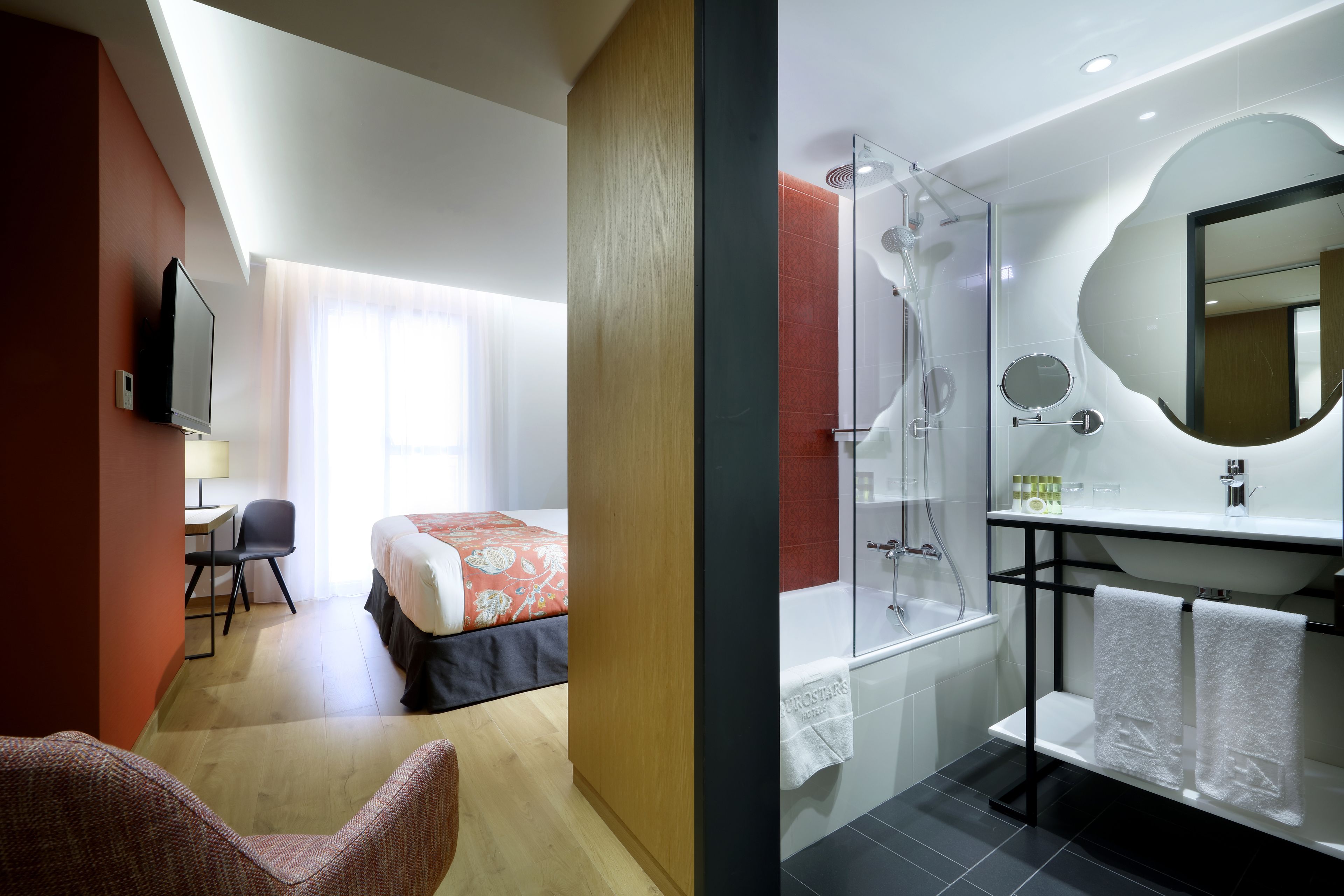 A hotel room with a bed, desk, chair, and a bathroom with a shower, sink, and mirror.
