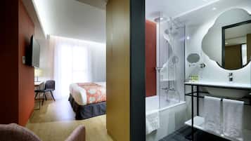 Una habitación de hotel con cama, escritorio, silla y baño completo con ducha, lavamanos y espejo.
