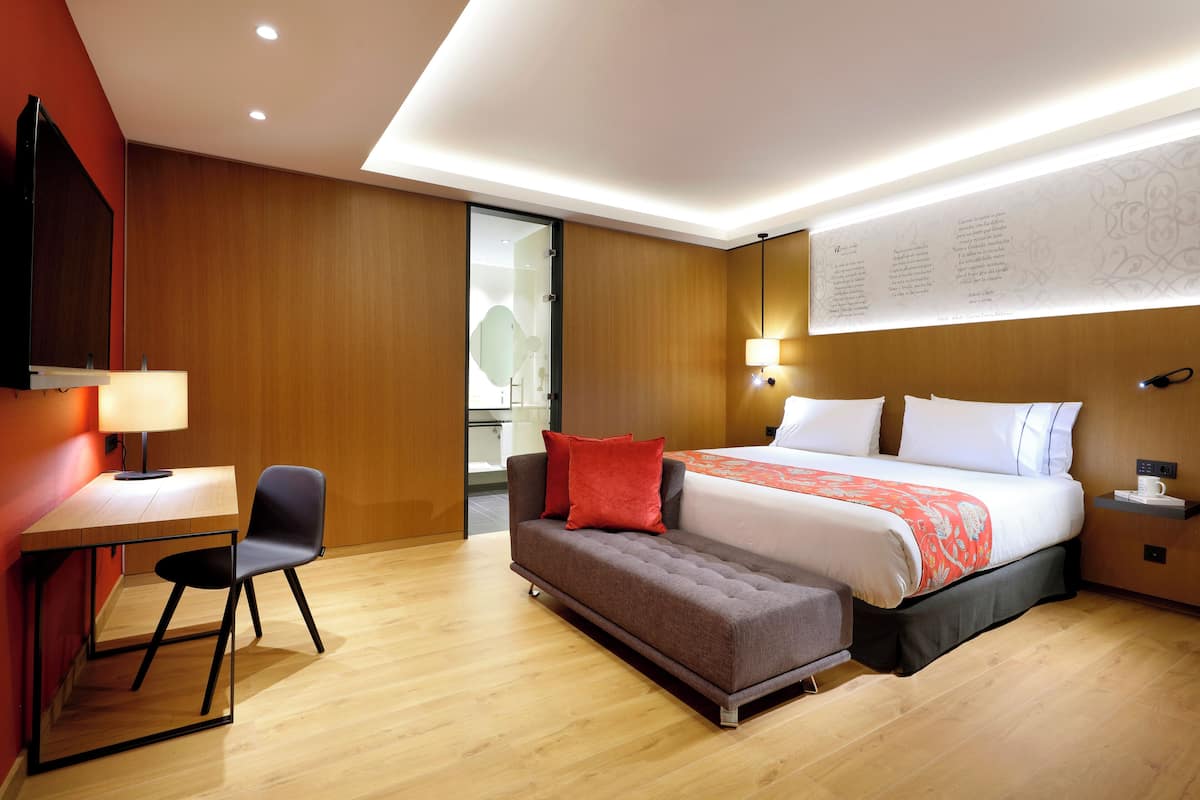Ein modernes Hotelzimmer mit Holzboden, einem Bett mit weißer und orangefarbener Bettwäsche, einem Schreibtisch mit Stuhl und einem an der Wand befestigten Fernseher.