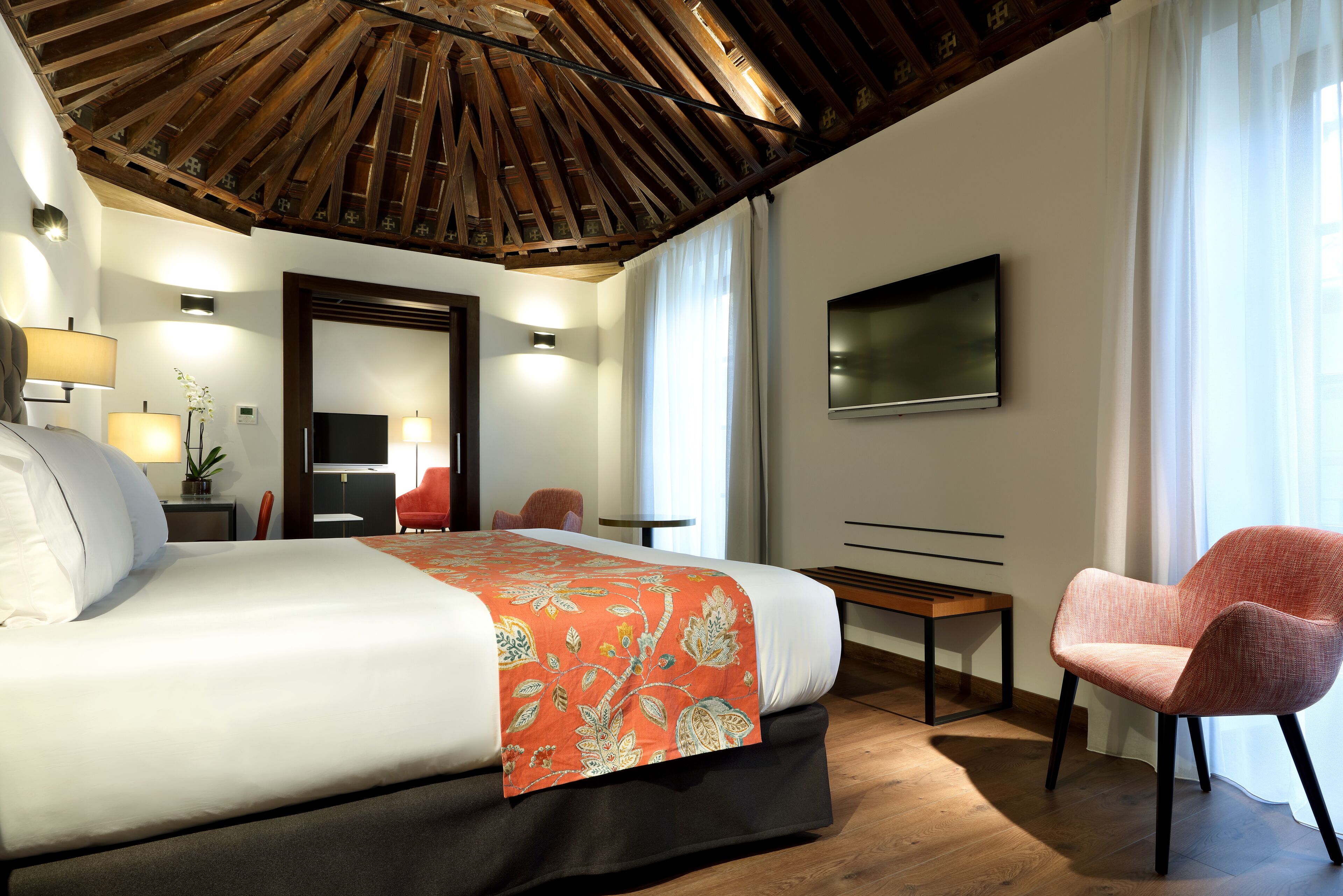 Habitación de hotel moderna con una cama grande, techo de madera, televisor de pantalla plana, una silla roja y una pequeña mesa.