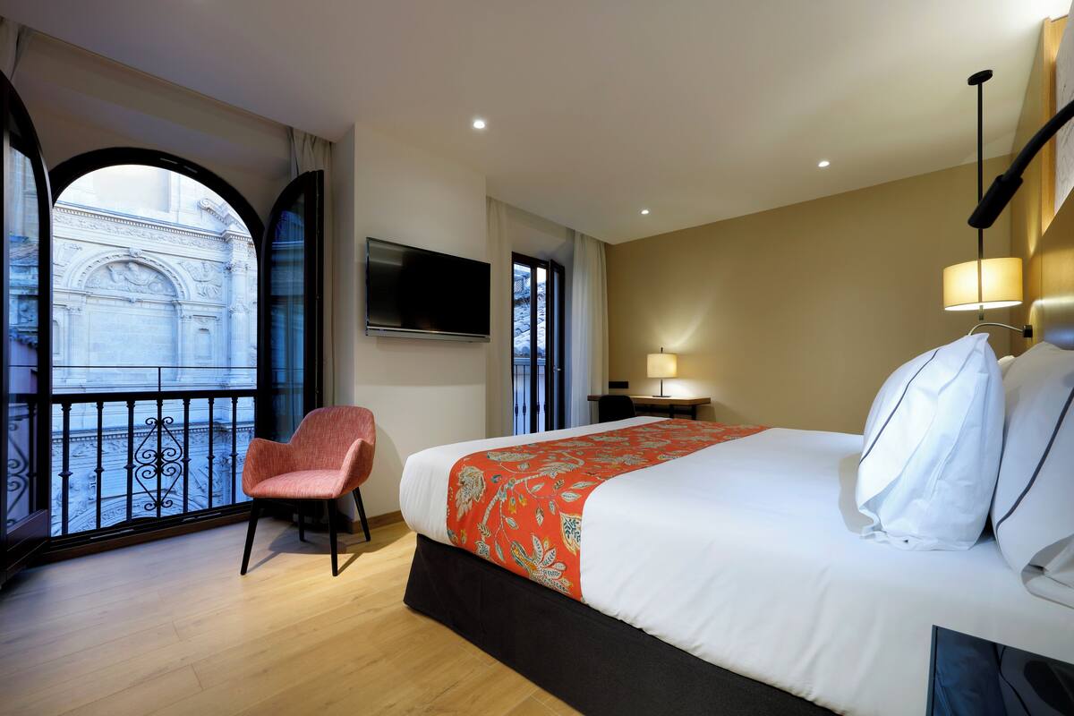 Ein Hotelzimmer mit einem großen Bett, einem Sessel, einem Fernseher und Blick auf ein Gebäude vom Balkon aus.
