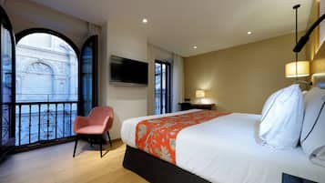 Habitación de hotel con una cama grande, una silla, un televisor y vistas a un edificio desde un balcón.