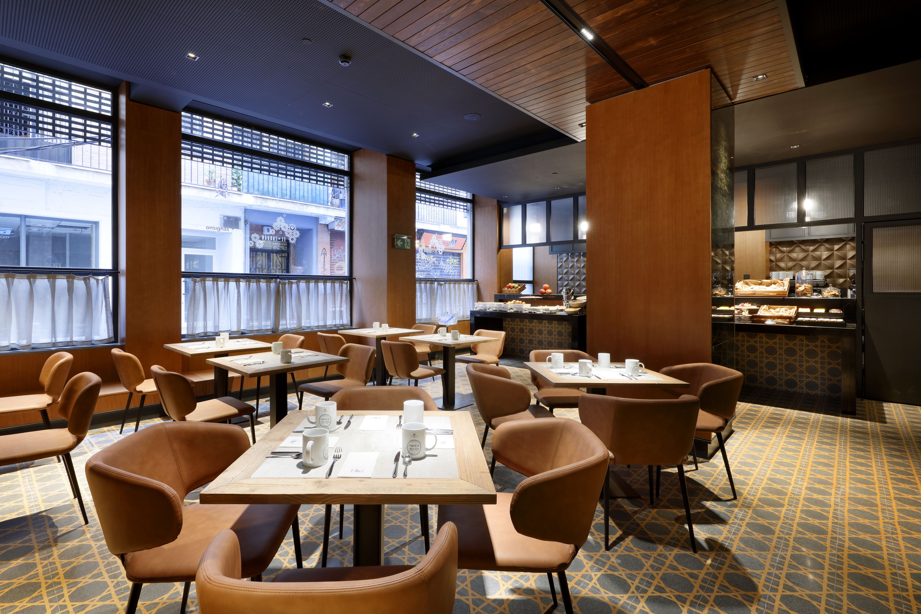 Un restaurante con mesas, sillas, una zona de cocina y amplios ventanales con vistas a la calle.