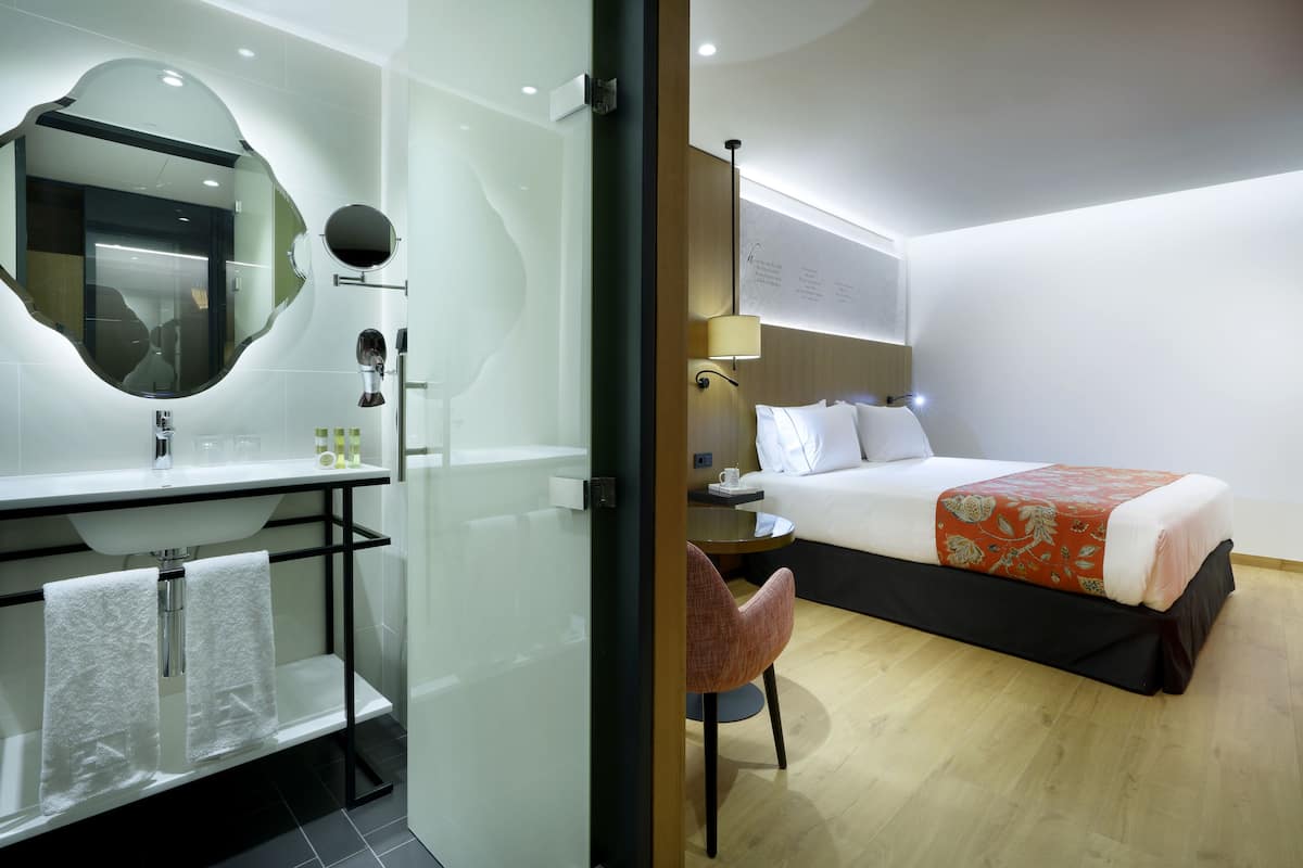 Ein modernes Hotelzimmer mit Duschkabine aus Glas, einem runden Spiegel, einem Nachttisch mit Lampe, einem Bett mit Blumenmuster-Bettdecke und einem Sessel.
