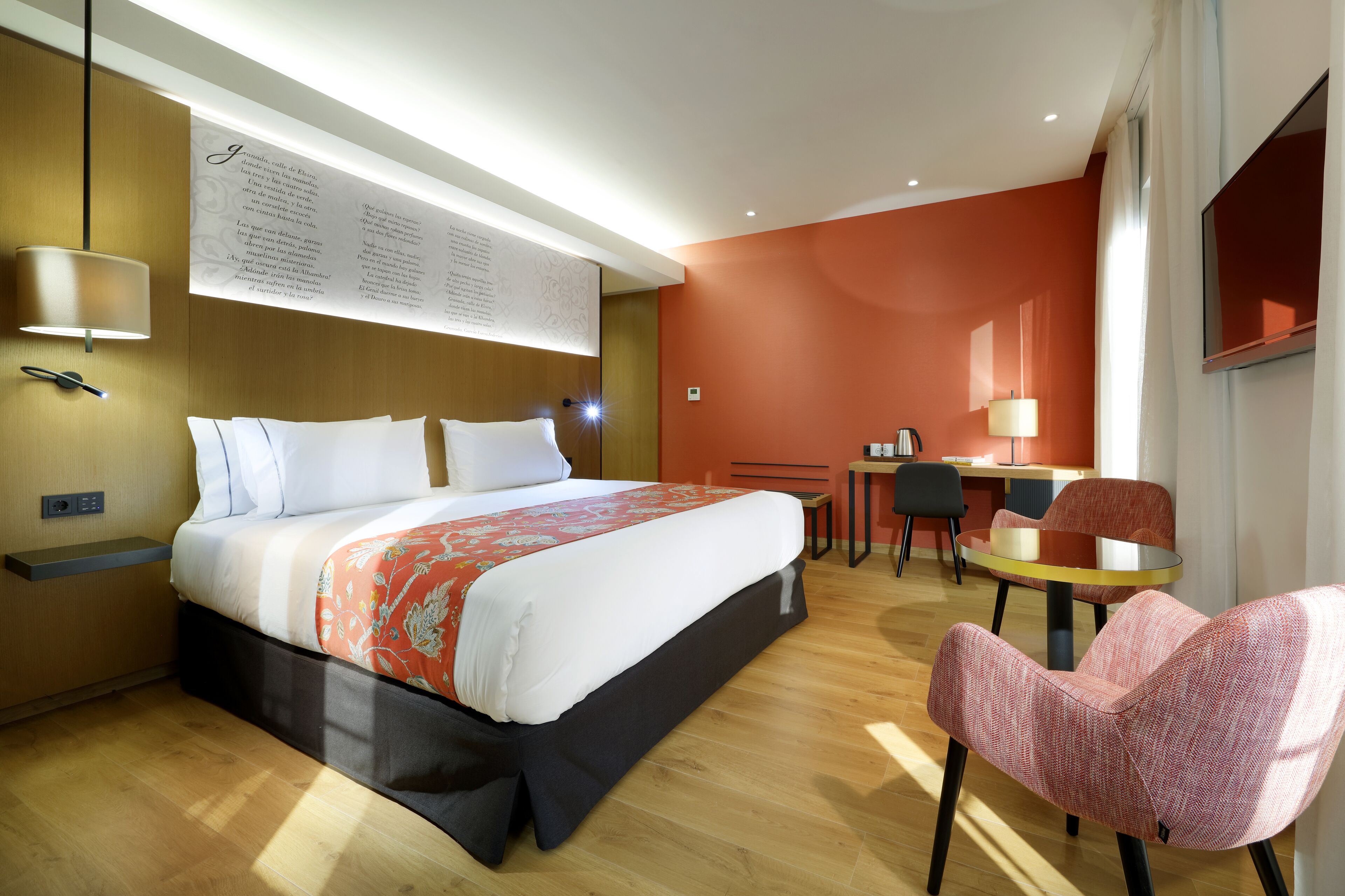 A hotel room with a bed, a desk, a chair, and a wall-mounted lamp.