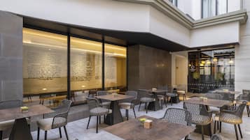Un área de comedor al aire libre con varias mesas y sillas, un muro de cristal con texto y la fachada de un edificio moderno.