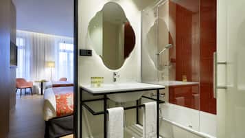 Una habitación de hotel moderna con una cama grande, un baño con ducha de vidrio y una zona de estar con una silla y una lámpara.
