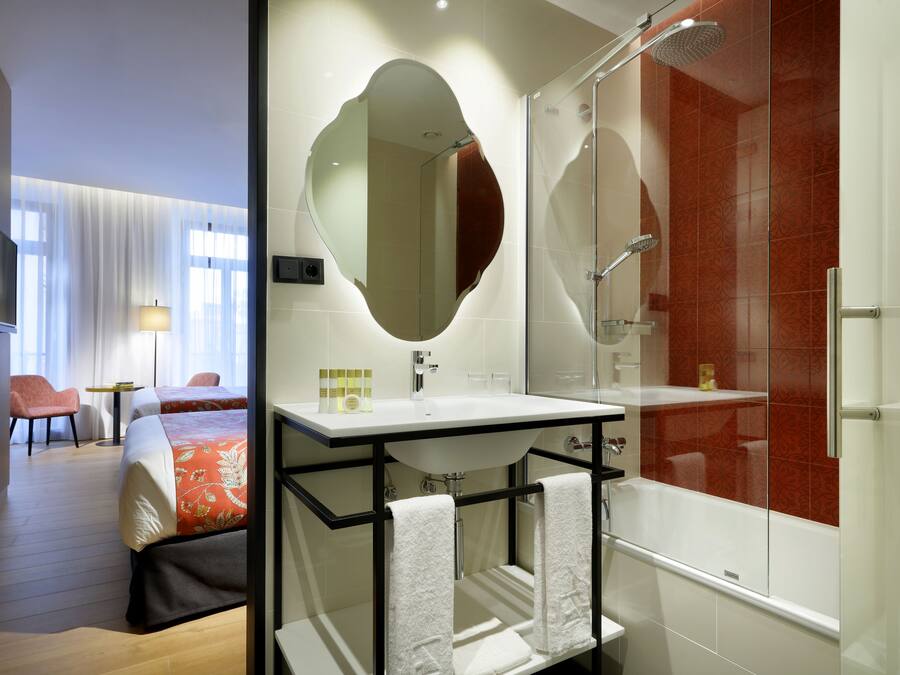 Una habitación de hotel moderna con una cama grande, un baño con ducha de vidrio y una zona de estar con una silla y una lámpara.