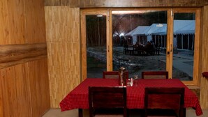 A dining area with a table set for four, wooden paneled walls, and a view of an outdoor seating area through glass doors. - Ladakh Tarrain Camp (Leh)