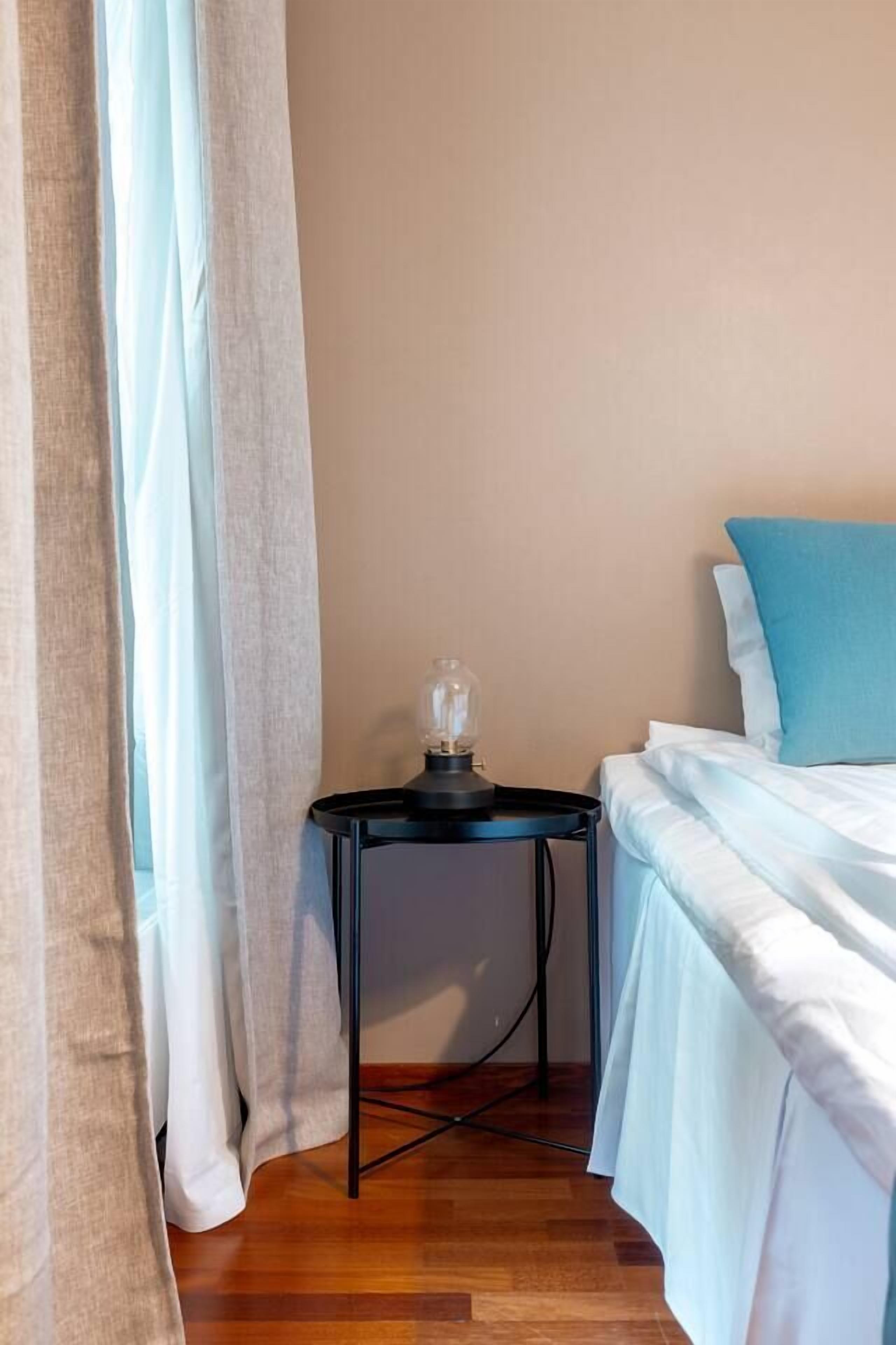 Ein Schlafzimmer mit einem Bett, einem Nachttisch, einer Lampe und einem Fenster mit Vorhängen.
