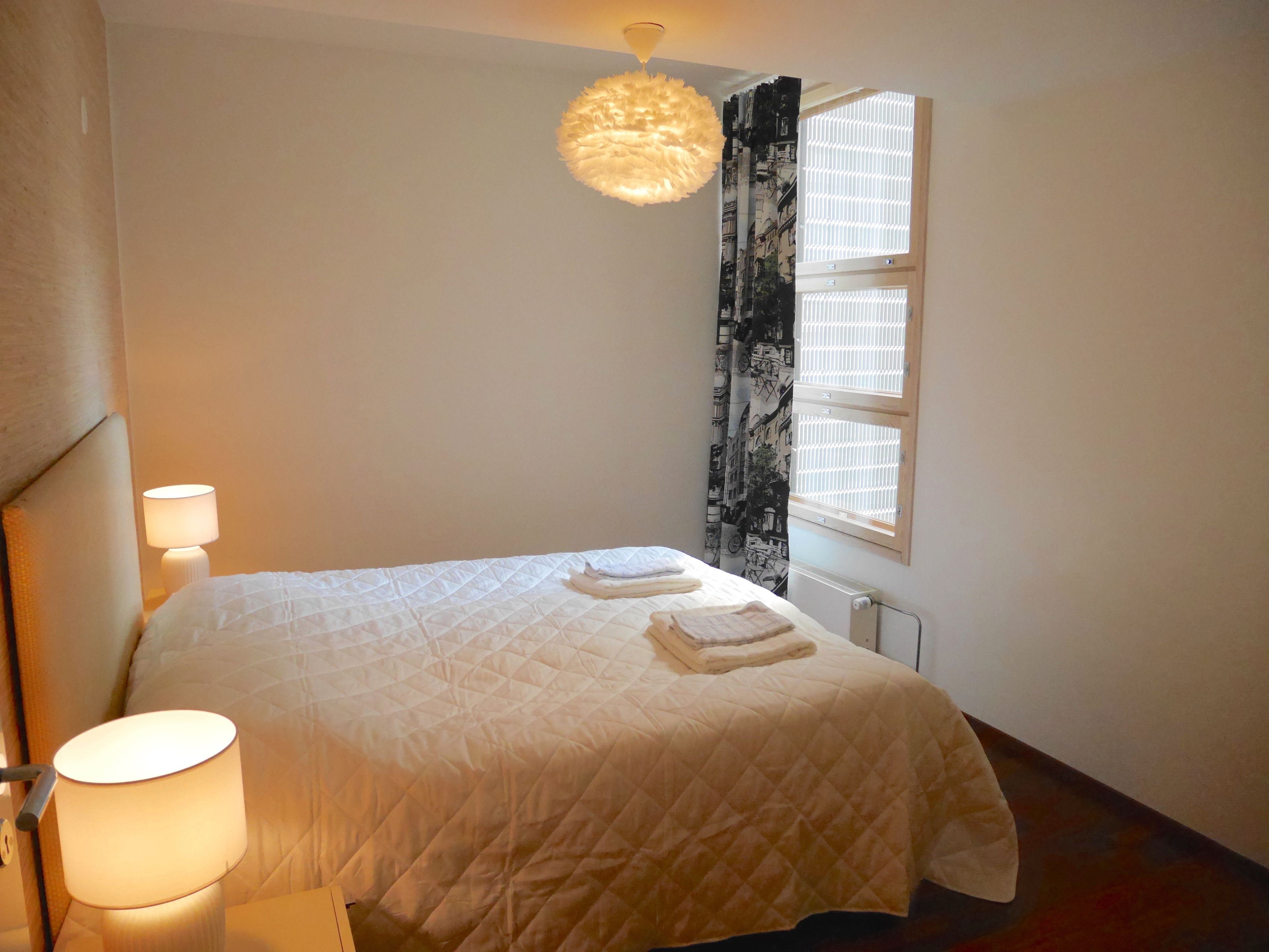 A neatly made bed with a white quilt, a bedside lamp, and a window with blinds.