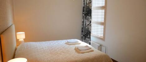 A neatly made bed with a white quilt, a bedside lamp, and a window with blinds.